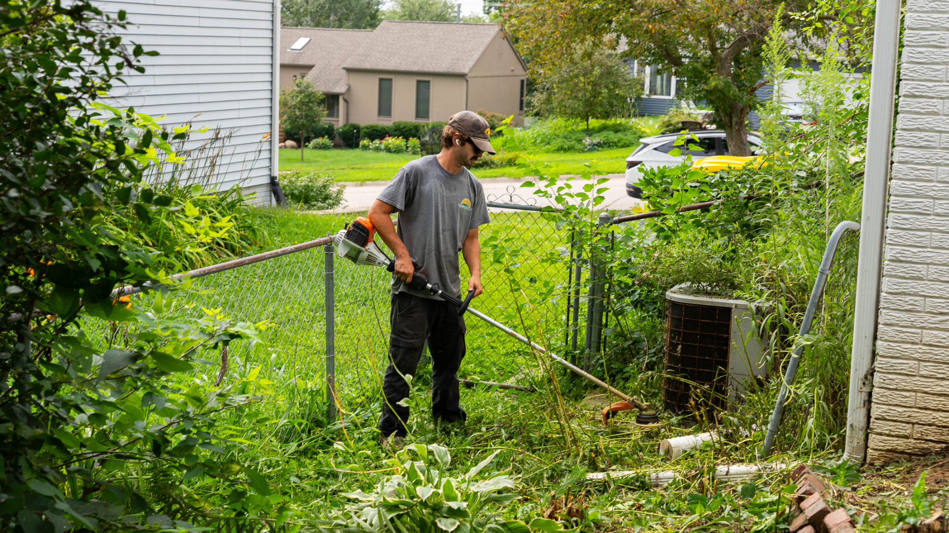 Heartland Grounds crew member string trimming an overgrown yard and fence line